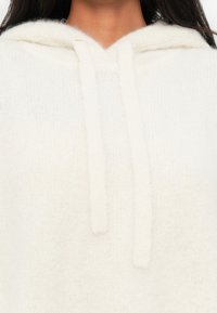 Hoodie bianco e morbido realizzato in materiale peloso con cappuccio regolabile. Presenta una finitura testurizzata che crea un aspetto accogliente.