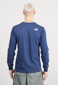 Marinblå långärmad t-shirt från The North Face med rundad hals, som har en liten logotyp på övre delen av ryggen och randiga detaljer på ärmarna.