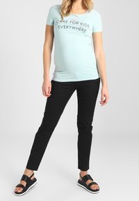 T-shirt bleu clair avec le texte "PRENEZ SOIN DES ENFANTS PARTOUT", pantalon skinny noir et sandales plates noires avec semelles blanches.