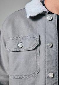 Grå denimjakke med en hvid shearlingkrave, trykknaplukning og en brystlomme med et særpræget mønster og pæne sting.