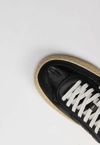 Sneaker in pelle nera con lacci bianchi, punta rotonda, parte superiore testurizzata e suola in gomma beige con un design intrecciato.