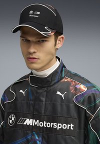 Schwarze Kappe mit weißen Akzenten und BMW Motorsport-Logo, kombiniert mit einer schwarzen Rennjacke mit bunten Mustern und Puma-Logos.
