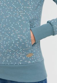 Sudadera azul claro con un patrón moteado y círculos en tonos pastel. Incluye un bolsillo lateral y puños y dobladillo acanalados. Tejido duradero con etiqueta de logo.