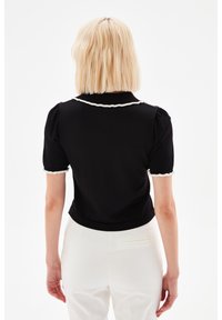 Black short-sleeve top with a white ruffled collar and cuffs, featuring a fitted design and smooth texture. Background is solid white.