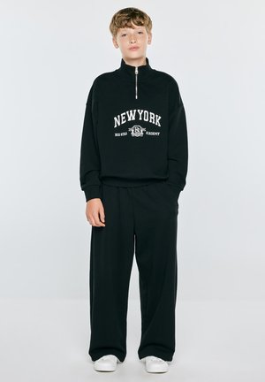 Młoda osoba w czarnej bluzie z kapturem New York z zamkiem błyskawicznym, czarnych szerokich spodniach i białych trampkach stoi na jednolitym tle.
