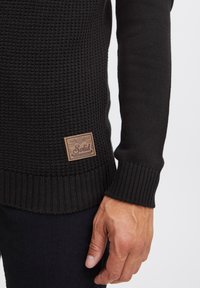 Knitted black sweater featuring a textured pattern, ribbed cuffs, and a brown leather label on the lower side with brand details.