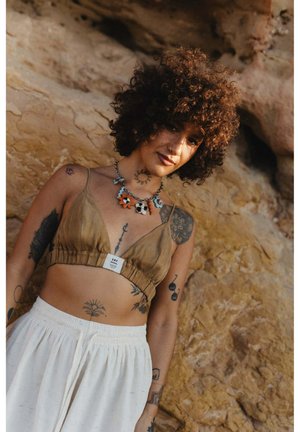Kobieta o kręconych włosach, ubrana w beżowy krótki top i białą spódnicę, prezentująca liczne tatuaże oraz kolorowy naszyjnik w kształcie kwiatów na tle skalistego krajobrazu.