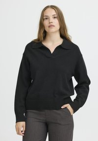 Juodas megztas svarkas su V formos iškirpte ir apykakle. Turi ilgus rankovės ir įlanką. Nešiojamas su pilkomis kelnėmis.