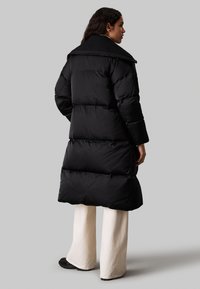 Abrigo largo negro tipo plumas con diseño acolchado, cuello alto y bolsillos laterales. Lleva puesto sobre pantalones anchos color crema y zapatos negros. Textura suave.