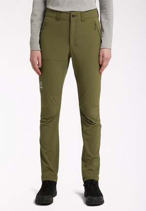 Personne portant un pantalon de randonnée vert olive avec des poches zippées et des chaussures de randonnée noires, debout sur un fond blanc.