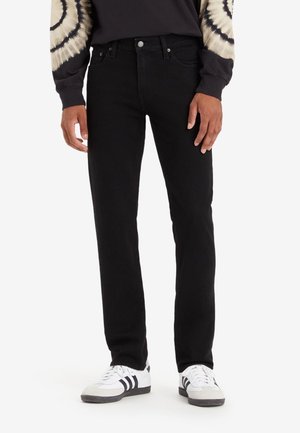 Mann trägt schwarze Jeans mit geradem Bein, ein schwarz-beiges Tie-Dye-Langarmshirt und weiße Sneakers mit schwarzen Streifen.