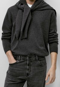Maglione grigio di lana con cappuccio e sciarpa drappeggiata. Paio di jeans grigio scuro con dettagli di bottoni e tasche. Texture liscia.