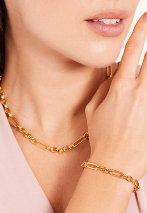 Goldkette und Armband mit länglichen Gliedern; glatte Textur mit glänzender Oberfläche; getragen zu einem hellrosa Oberteil.
