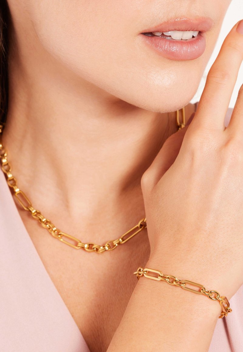 Goldkette und Armband mit länglichen Gliedern; glatte Textur mit glänzender Oberfläche; getragen zu einem hellrosa Oberteil.