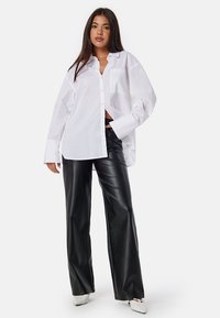 Witte oversized button-up blouse met lange mouwen, gecombineerd met zwarte hoogsluitende nep leren broek en witte puntige hakken.