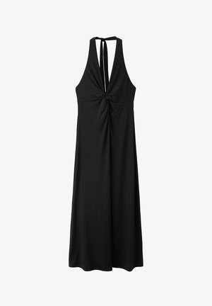 Femme aux cheveux bruns courts portant une robe noire dos nu à col halter et des sandales noires, debout devant un fond clair uni.