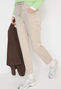 Personne portant un pantalon beige, des chaussettes blanches, des baskets argentées, un pull vert ; tenant une veste marron texturée dans la main gauche.
