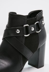 Czarny botek aż kostkę wykonany z gładkiej i zamszowej skóry, z szerokim paskiem z metalowymi elementami w kolorze srebra i grubym obcasem.