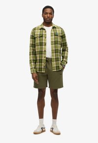Camisa de franela a cuadros en verde y amarillo, camiseta blanca de cuello redondo, pantalones cortos verde oliva, zapatillas blancas con detalles en negro y calcetines cortos.