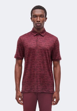 Homme portant un polo bordeaux à manches courtes avec un motif de couronne, debout les bras détendus devant un fond clair uni.