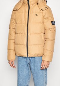 Beige pufferjakke med høj krave, sort lynlås og logo patch på venstre ærme. Har trykknapper på skuldrene.