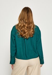 Tyrkysová košeľa s dlhými rukávmi a voľným strihom, s manžetami na gombíky a textúrovanou látkou. Nosí sa s béžovými nohavicami, čo vytvára ležérny štýl.