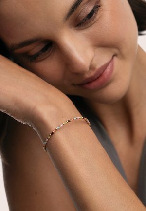 Pulsera de tono dorado con pequeñas cuentas multicolores en tonos pastel, llevada en la muñeca. El fondo es suave y neutro.