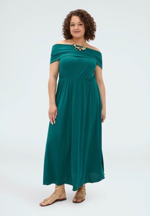 Kvinde med krøllet hår iført en teal off-shoulder maxikjole, guldfarvet statement-halskæde og gyldne sandaler med flade remme, stående på en hvid baggrund.