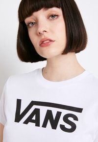 Bílá bavlněná tričko s velkým černým logem "VANS" v tučném, moderním písmu. Kulatý výstřih a krátké rukávy. Hladký povrch.