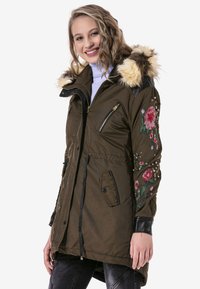 Olivgrüne Parka mit Kapuze aus Kunstfell, bestickten Blumenmotiven auf den Ärmeln und mit Reißverschluss versehenen Taschen.