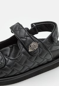 Czarna skórzana sandał z pikowanym wzorem, regulowanym paskiem i metalowymi akcentami. Posiada wyściełaną wkładkę oraz teksturowaną podeszwę.