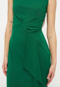 Κοντινό πλάνο ενός πράσινου φορέματος με στρώσεις βολάν στη μέση, φορεμένο από ένα άτομο με μικρά τατουάζ και στα δύο αντιβράχια.