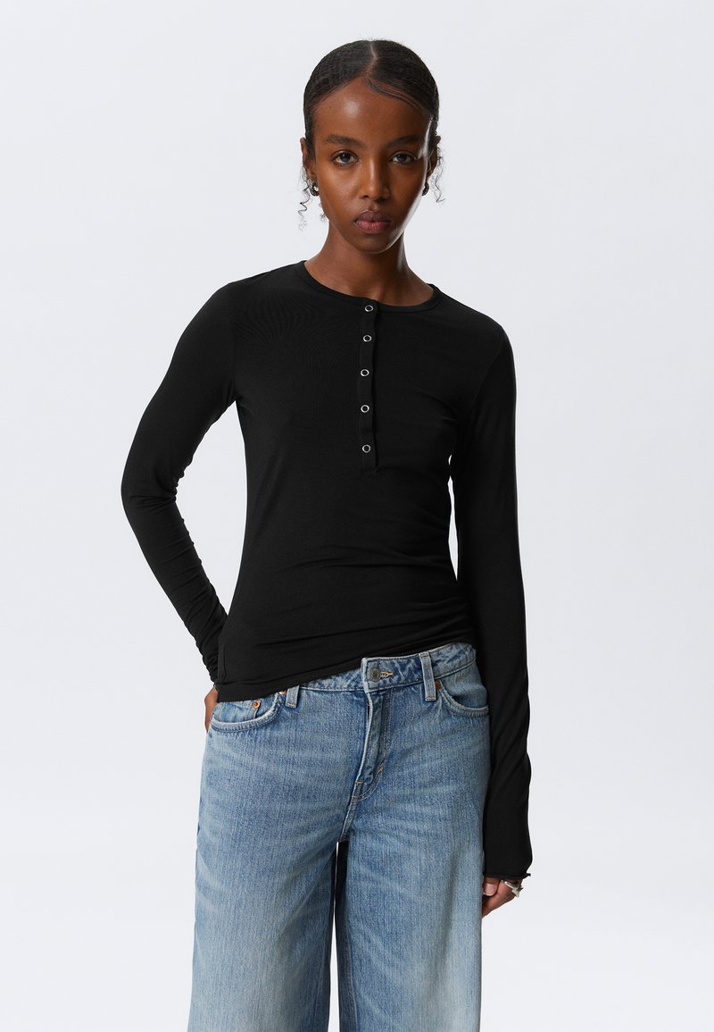 Svart langermet skjorte med rund hals og fem knapper foran, sammen med lyseblå vide jeans. Glatt tekstur.