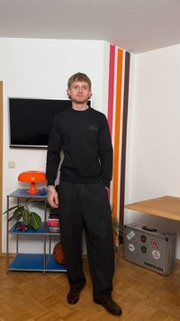 Młody mężczyzna w czarnym stroju stoi w pokoju z paskowaną ścianą, pomarańczową lampą, piłką do koszykówki, walizką oraz małą półką z roślinami i książkami.