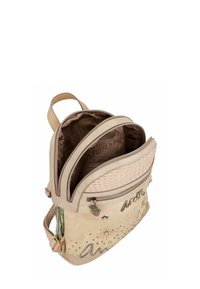 Sac à dos beige avec une surface texturée, comportant plusieurs compartiments zippés, des coutures décoratives et un pendentif décoratif sur un côté.