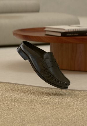 Μαύρο δερμάτινο μοκασίνι κρεμασμένο πάνω από ένα μπεζ υφαντό χαλί, στο φόντο στρογγυλό ξύλινο τραπέζι και βιβλία.