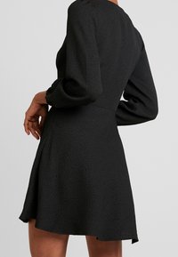 Abito nero con tessuto strutturato, maniche lunghe, vita definita e gonna svasata, visto da dietro.