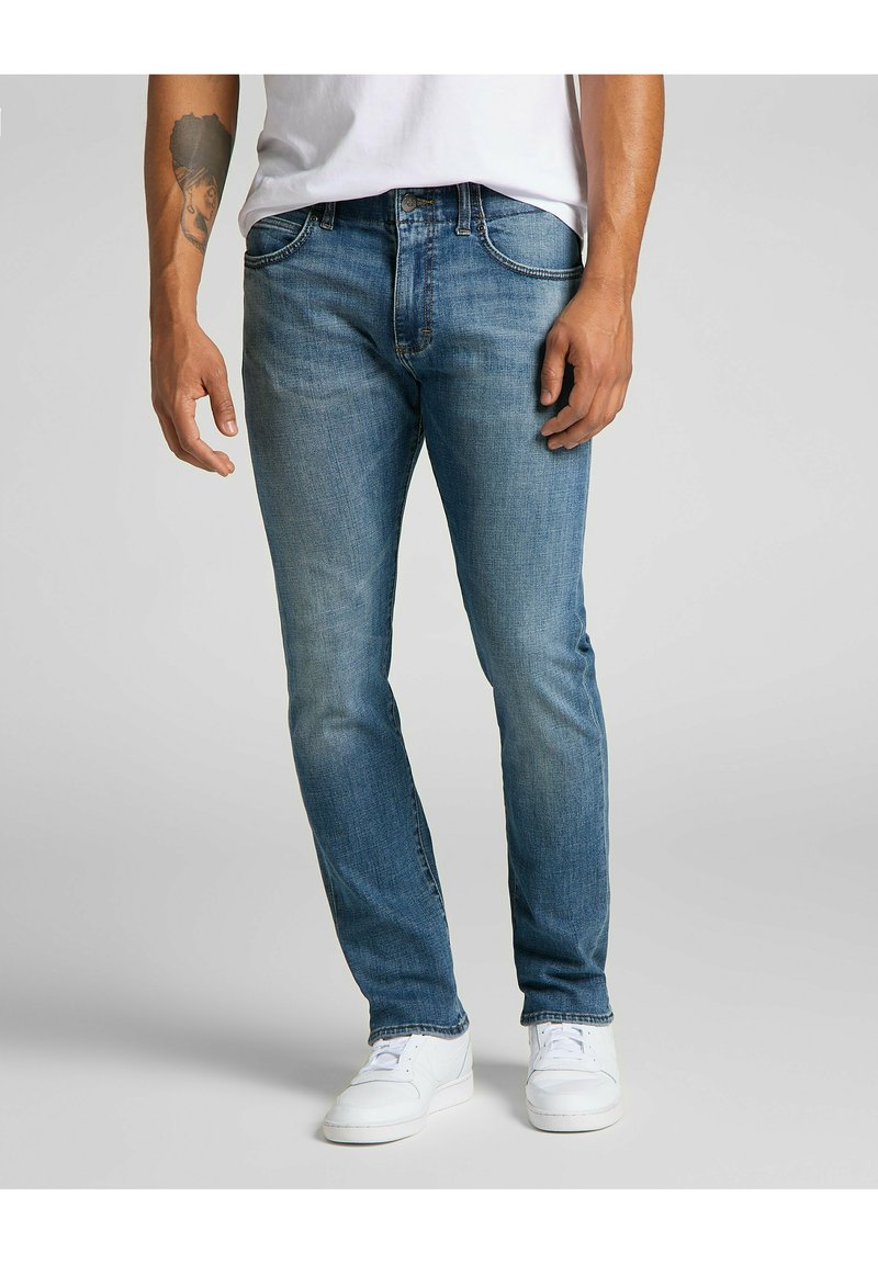 Jeans en denim bleu clair avec une coupe slim, cinq poches, un léger dégradé et des coutures contrastées, associés à des baskets blanches.