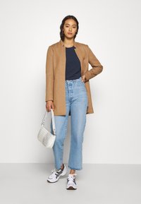 Beige, strukturert jakke laget av mykt materiale, kombinert med en marineblå topp og lyseblå høyt liv jeans. Hvit teksturert håndveske og sporty sko.