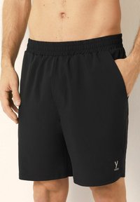 Mannelijke torso in zwarte sportshorts met elastische tailleband en zakken, staande tegen een neutrale achtergrond met een hand in de zak.