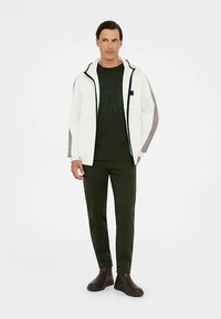 Weißer Hoodie mit Reißverschluss, graue Seitenpaneele, dunkelgrünes T-Shirt, olivgrüne Hose und braune Knöchelstiefel. Einfaches Design, neutrale Farben.