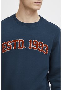 Sötétkék pulóver kerek nyakkal, domború, texturált narancssárga felirattal "ESTD. 1993" a mellkason. Puha pamut anyagból készült.