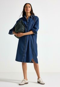 Jeansklänning i mörkblått med knäppning framtill, uppvikta ärmar och hög-låg nederkant. Modellen håller en grön väska och har vita sneakers på sig.