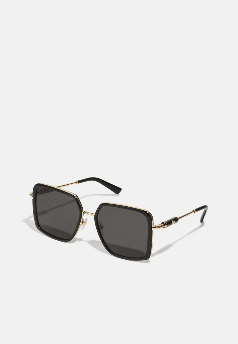 Gafas de sol con lentes cuadrados negros, montura de metal dorado y puntas de las patillas negras. Las características incluyen un puente delgado y herrajes decorativos.