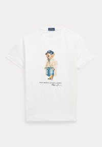 T-shirt blanc avec un graphique d’un ours portant une casquette NY, un pull, un jean et un manteau, avec le texte "POLO BEAR by RALPH LAUREN" en dessous.