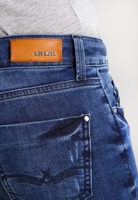 Džínové kalhoty v tmavě modré barvě s texturovaným povrchem. Mají koženou nášivku s nápisem "MOS MOSH" a viditelné švy na zadní kapse.