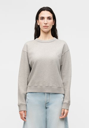 Moteris su ilgais tamsiais plaukais, vilkinti šviesiai pilką džemperį ir šviesiai mėlynas aukšto liemens džinsus, stovinti prieš paprastą baltą foną.