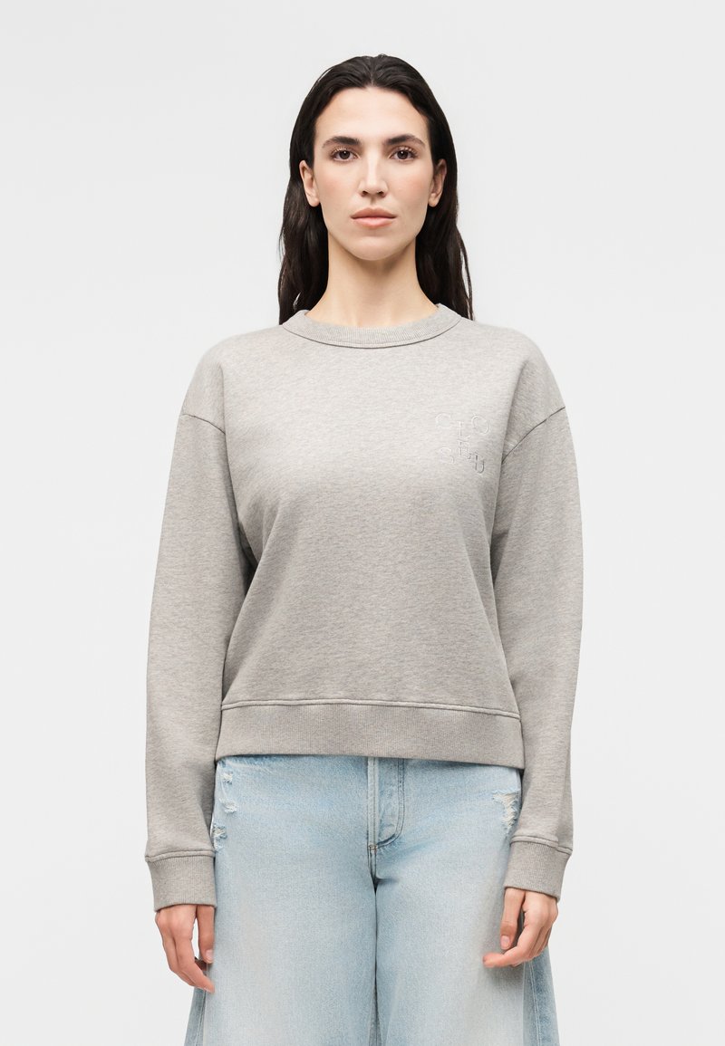 Γυναίκα με μακριά σκουρόχρωμα μαλλιά, που φοράει ανοιχτογρι ζακέτα και ανοιχτό μπλε ψηλόμεσα τζιν, στέκεται μπροστά σε απλό λευκό φόντο.