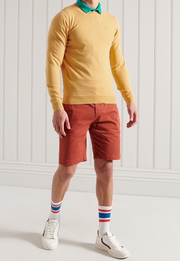 Pull en maille jaune avec un col vert, associé à un short rouge. Baskets blanches avec des chaussettes à rayures colorées. Tenue simple et décontractée.