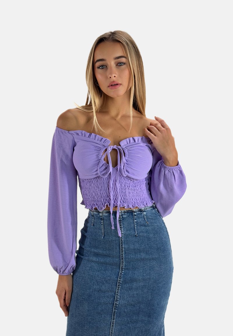 Blouse violette à épaules dénudées, cropped, avec un col à volants, un lien ajustable à l'avant et un corsage smocké. Associée à une jupe crayon en denim.